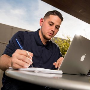 Photo of a male student sitting at a laptop and writing on a padlet of paper.